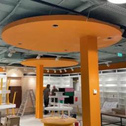 Wnętrze sklepu w trakcie wykończenia: pomarańczowe kolumny i okrągłe podwieszane elementy, widoczne regały, pracownicy i materiały budowlane.