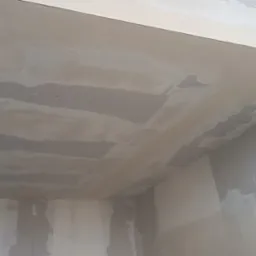 Wnętrze pokoju w trakcie prac wykończeniowych, widoczne ślady po szpachlowaniu na suficie i ścianach, częściowo pokryte białą farbą, ujęcie z dołu.