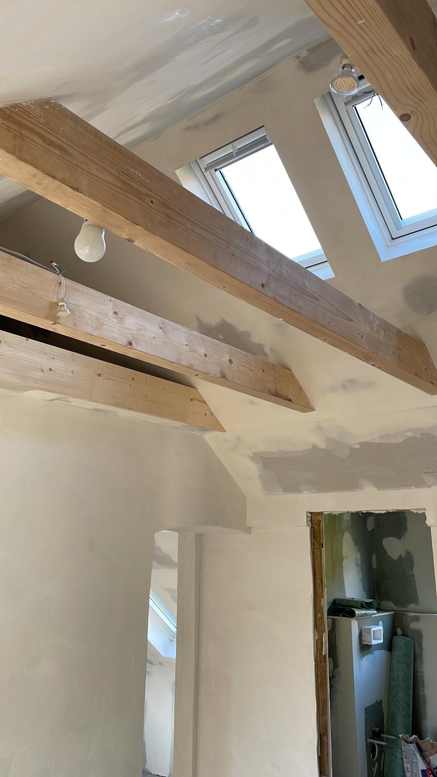 Wnętrze poddasza w trakcie wykańczania: ściany i sufit po szpachlowaniu, widoczne drewniane belki stropowe, okna dachowe i wisząca żarówka.