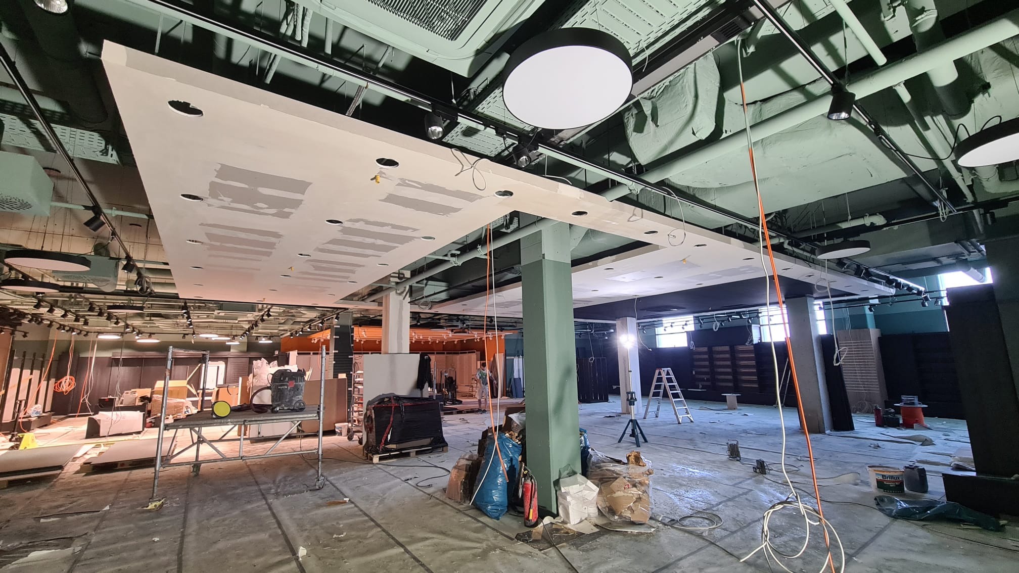 Sufit podwieszany w trakcie montażu w lokalu komercyjnym, widoczne szpachlowane łączenia płyt, instalacje pod sufitem oraz prowizoryczne oświetlenie budowlane.