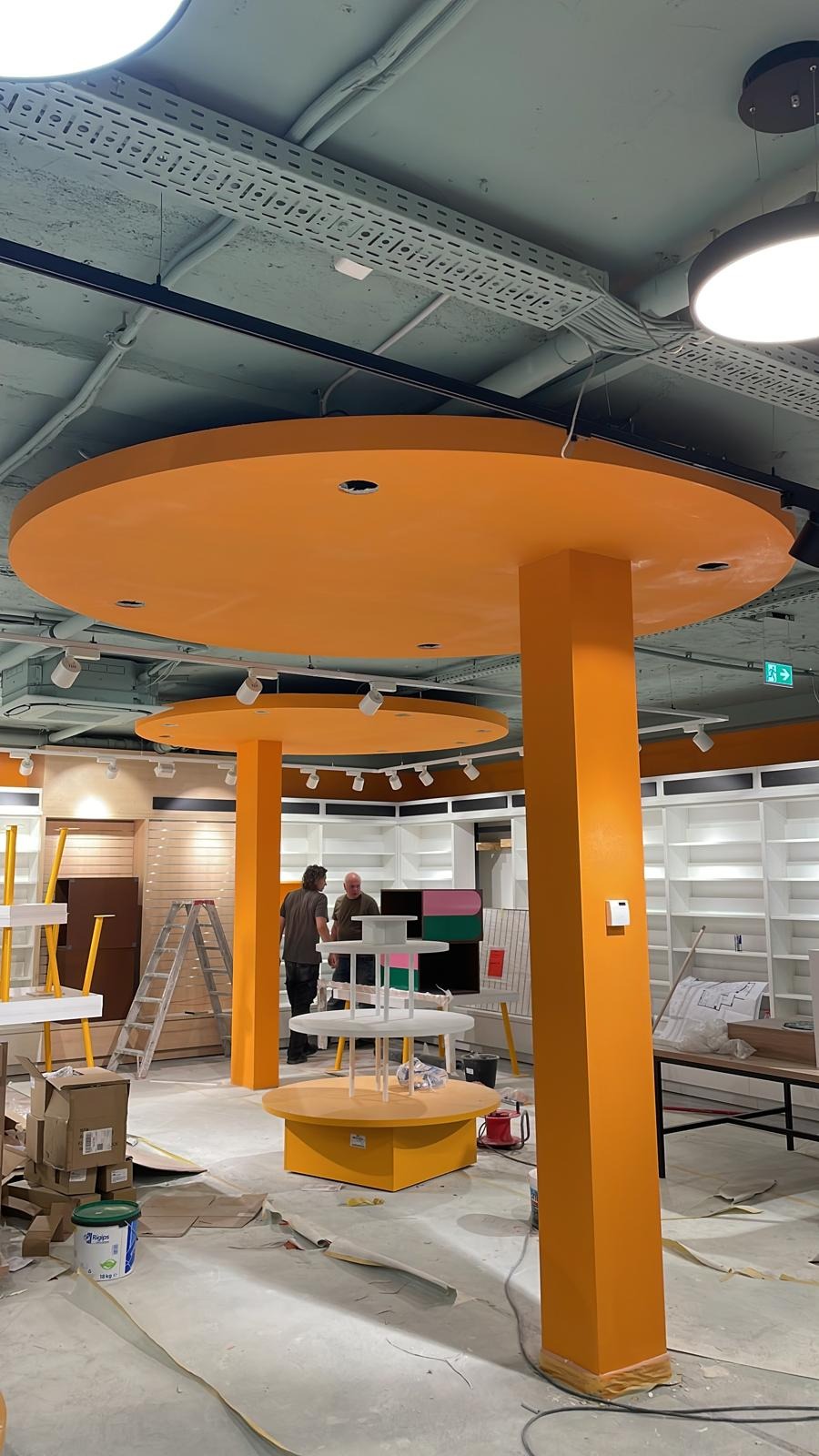 Wnętrze sklepu w trakcie wykończenia: pomarańczowe kolumny i okrągłe podwieszane elementy, widoczne regały, pracownicy i materiały budowlane.