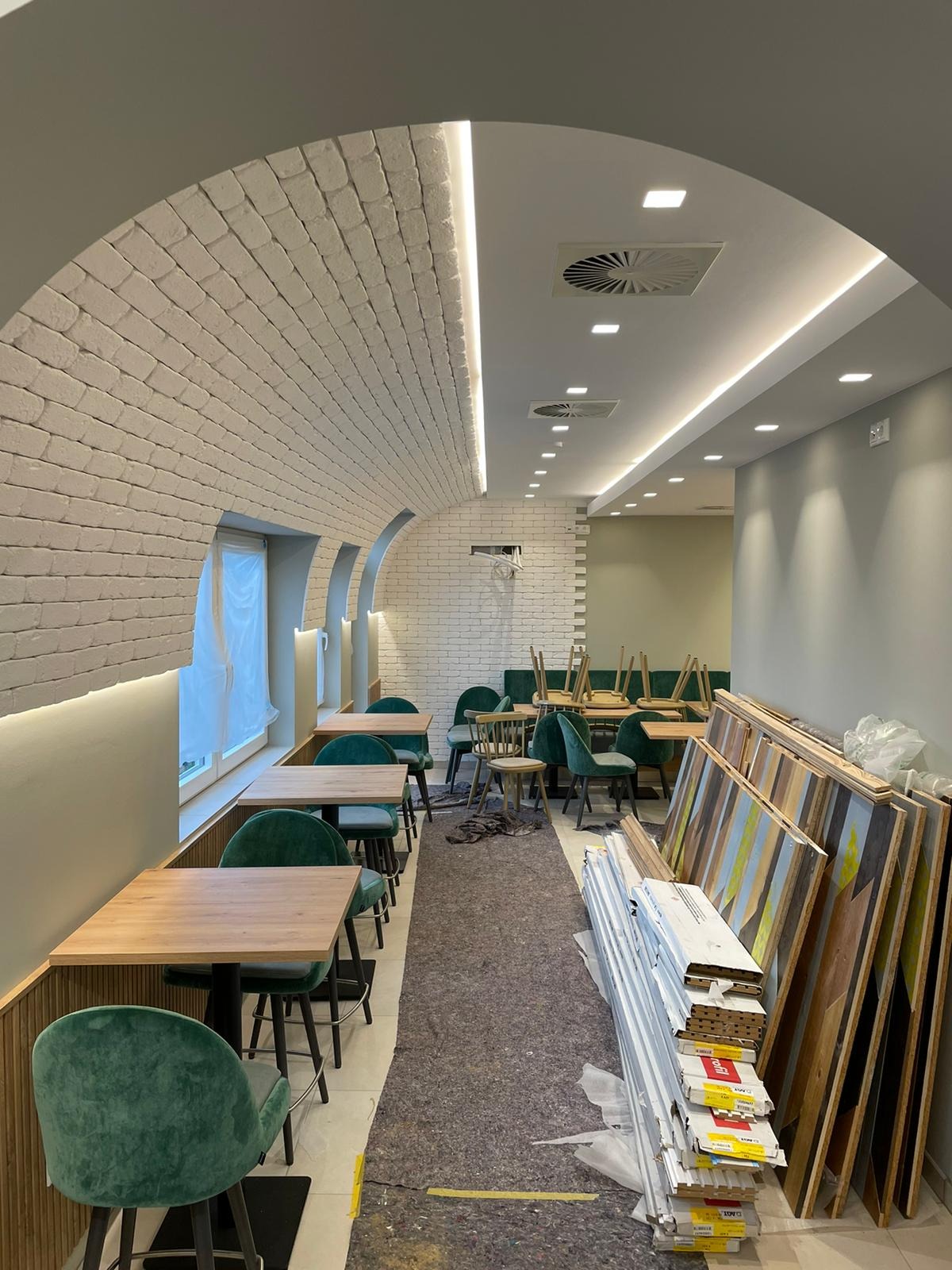 Wnętrze lokalu gastronomicznego w trakcie wykańczania: biała cegła na łukowym suficie, drewniane stoły z zielonymi krzesłami, zabezpieczona podłoga i materiały budowlane wzdłuż ściany.