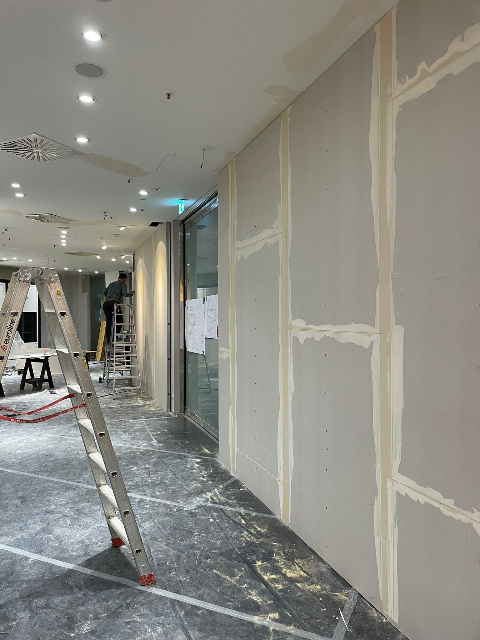 Wnętrze komercyjne w trakcie prac wykończeniowych: ściany z płyt gipsowo-kartonowych ze wstępnie nałożoną masą szpachlową na łączeniach, drabina aluminiowa z przodu, pracownik na drabinie w tle...