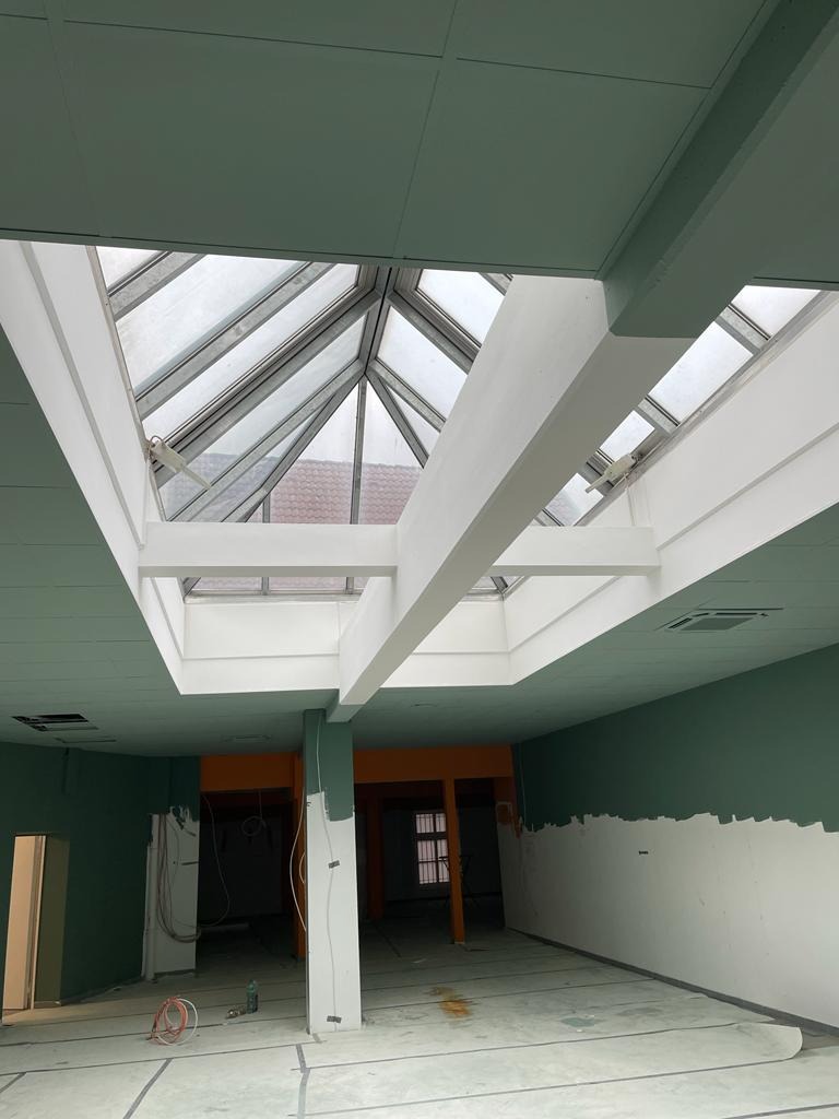 Wnętrze w trakcie prac wykończeniowych: sufit z zielonymi panelami, piramidalne okno dachowe z białymi ramami, ściany częściowo pomalowane na zielono, widoczne kable i niedomalowane fragmenty kolumn.