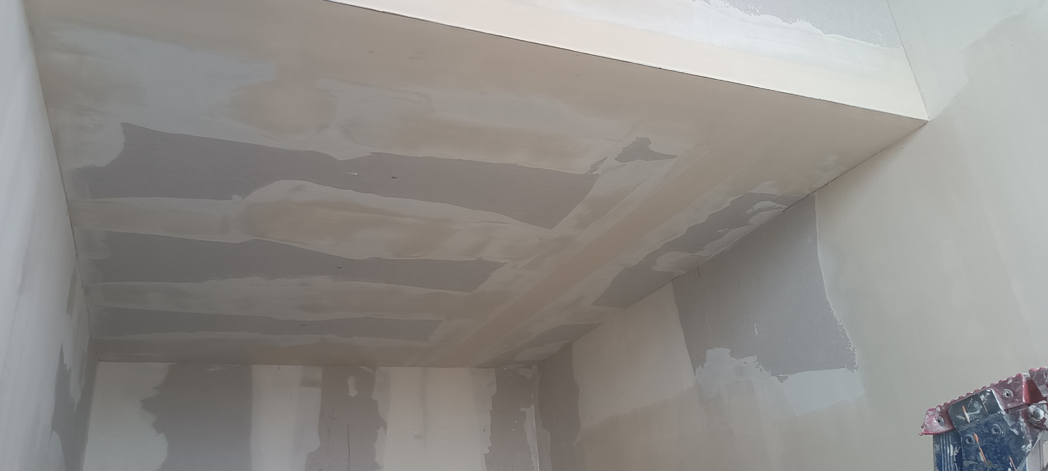 Wnętrze pokoju w trakcie prac wykończeniowych, widoczne ślady po szpachlowaniu na suficie i ścianach, częściowo pokryte białą farbą, ujęcie z dołu.