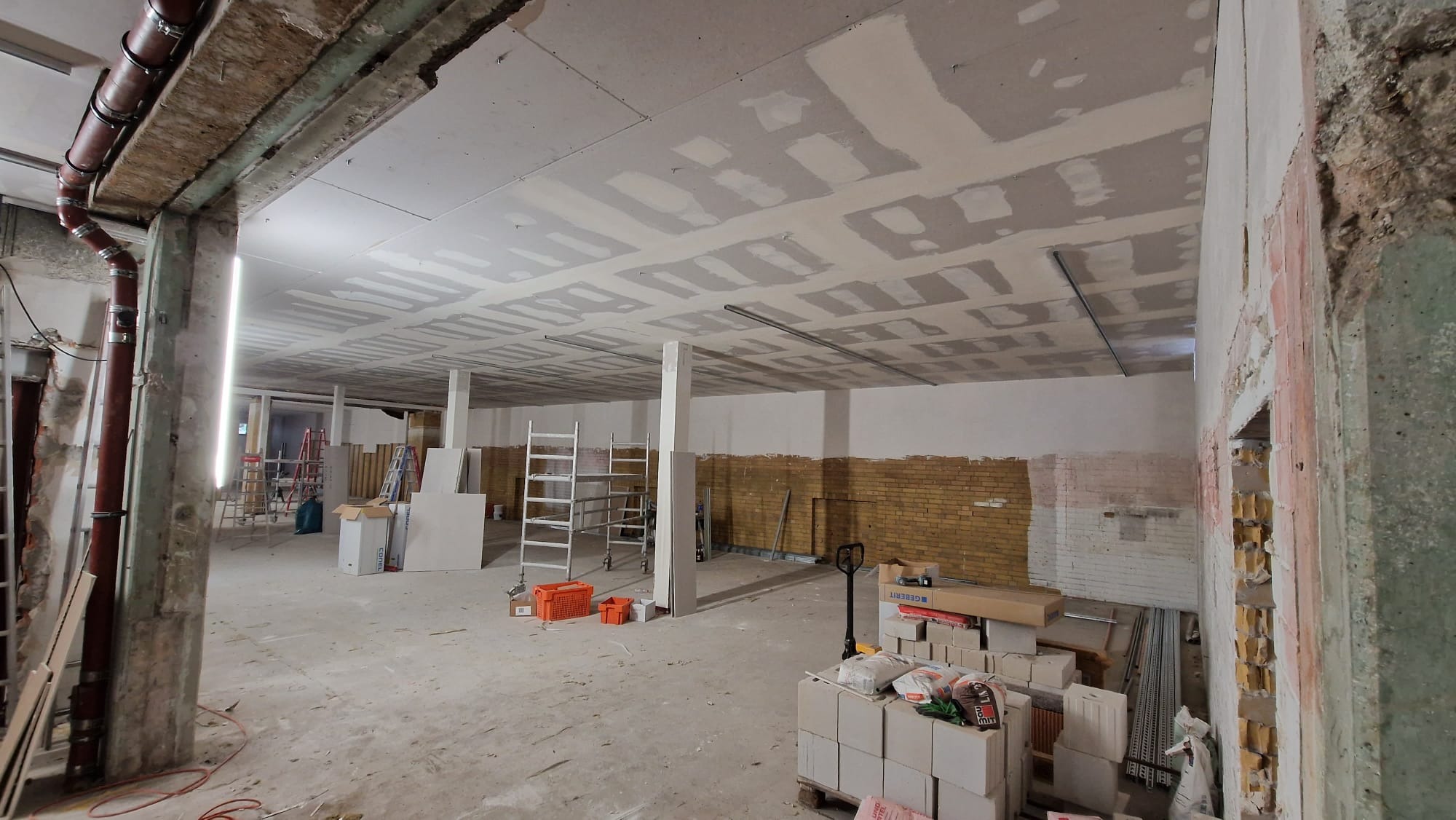 Wnętrze w trakcie remontu: sufit z płyt gipsowo-kartonowych po szpachlowaniu, fragmenty odsłoniętej cegły na ścianie, materiały budowlane i drabiny w przestrzeni.
