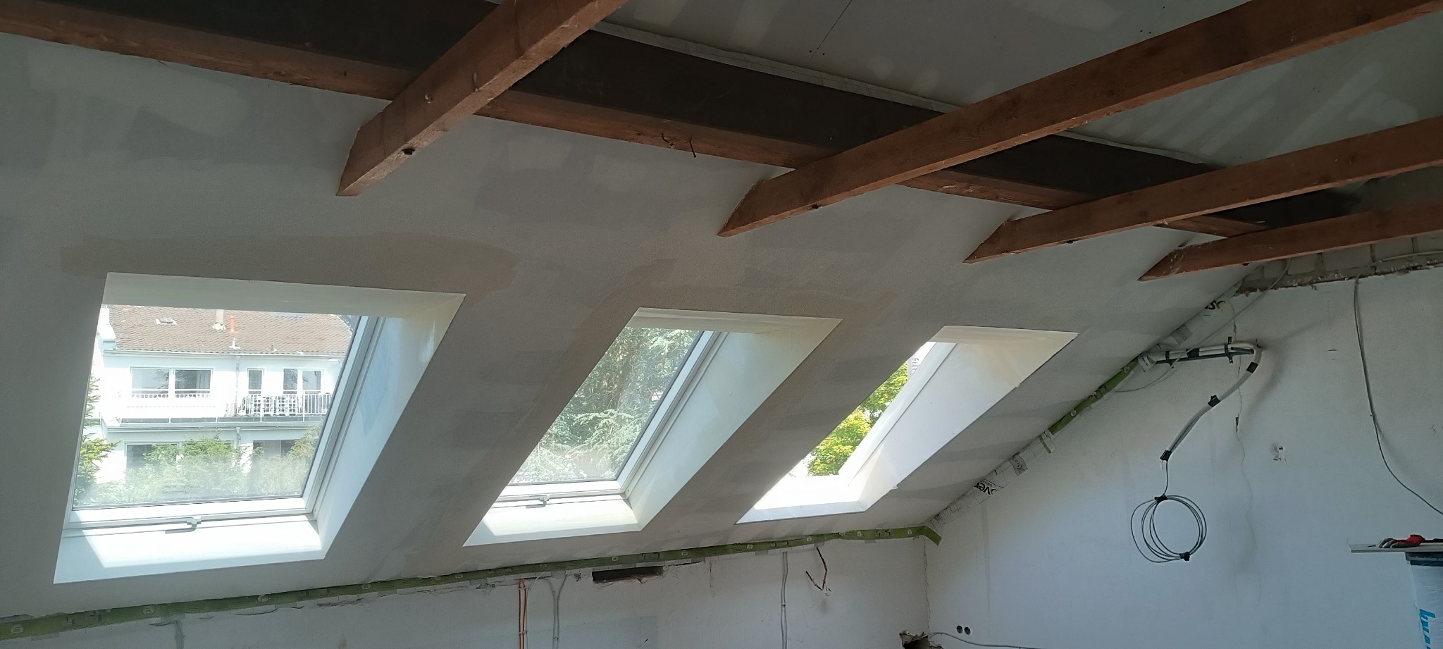 Poddasze w trakcie prac wykończeniowych, widoczne trzy okna dachowe, surowy tynk na ścianach i suficie, drewniane belki stropowe, instalacja elektryczna prowadzona po ścianie.
