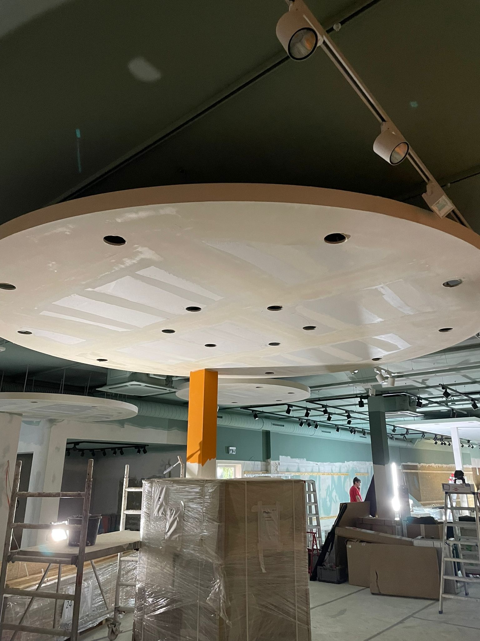 Niedokończony sufit podwieszany z okrągłymi elementami i otworami na oświetlenie, widoczne ślady szpachlowania, w tle rusztowania i zapakowane materiały budowlane.