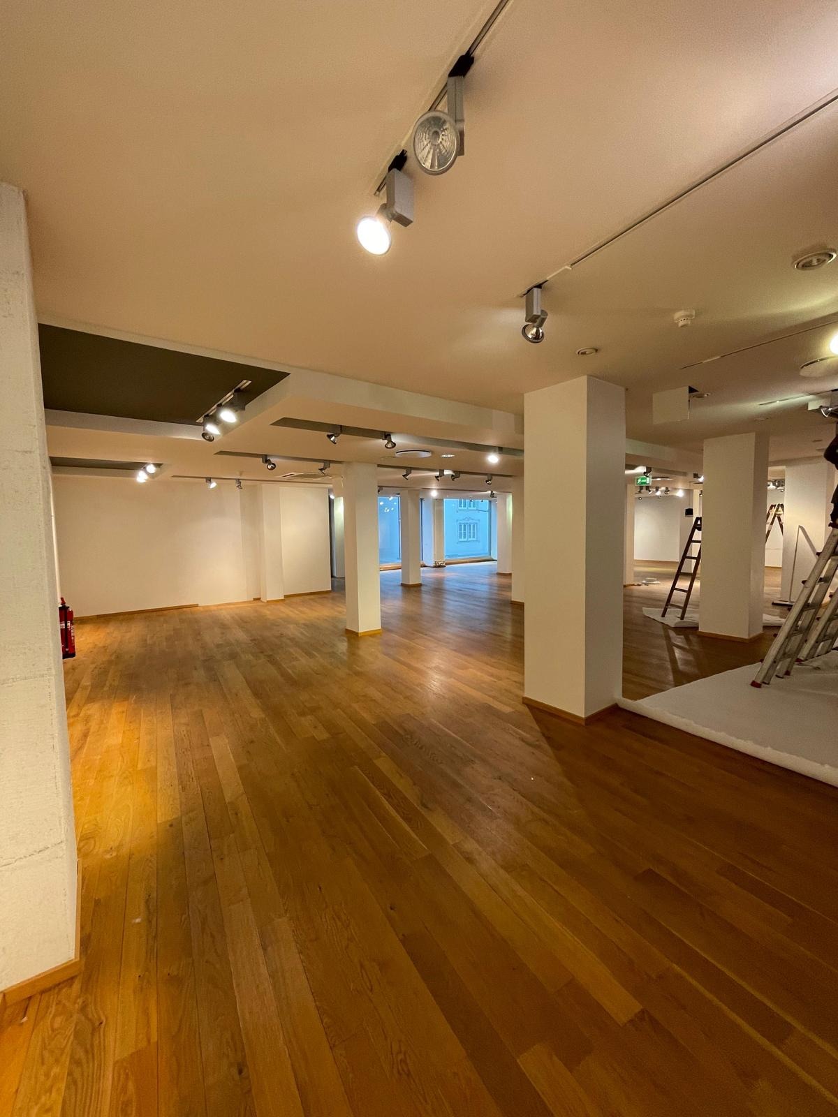 Przestronne wnętrze z drewnianą podłogą w trakcie prac wykończeniowych, widoczne drabiny i zabezpieczona podłoga, białe kolumny i oświetlenie punktowe.