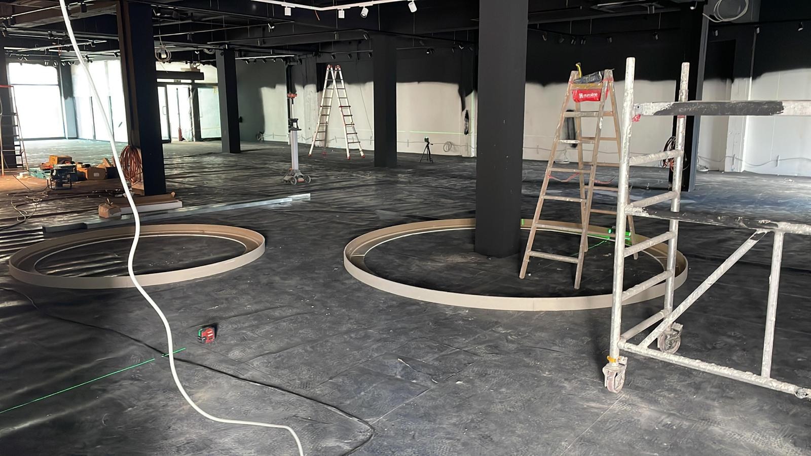 Wnętrze w trakcie prac wykończeniowych: przygotowanie do montażu okrągłych elementów dekoracyjnych sufitu podwieszanego, widoczne drabiny, rusztowanie i laserowy niwelator.