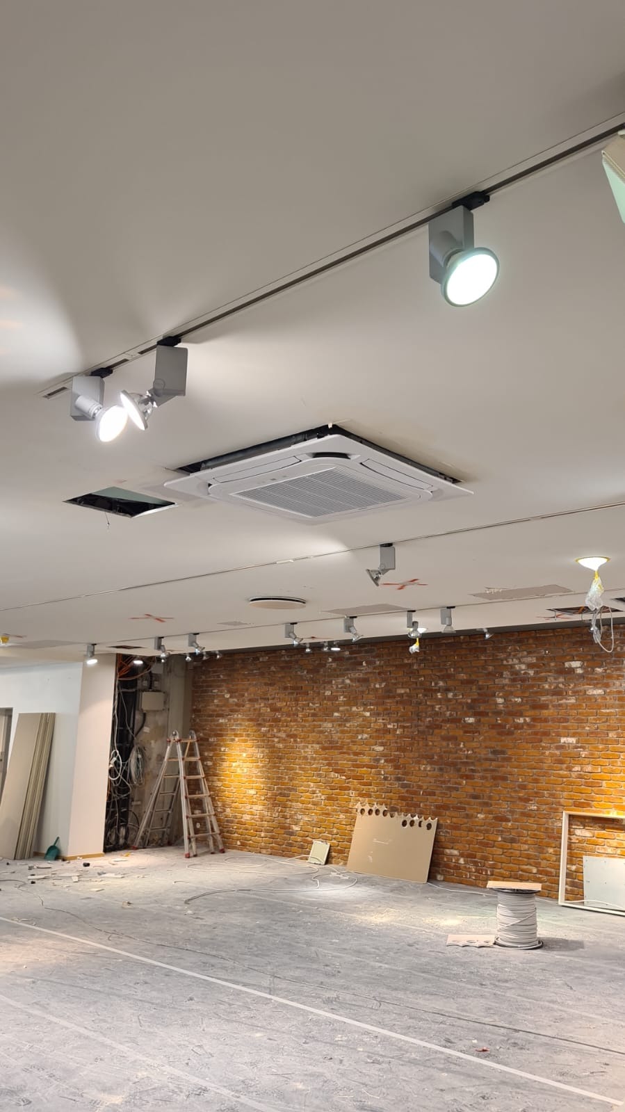 Wnętrze w trakcie prac wykończeniowych: widoczna ceglana ściana, sufit z zamontowanym systemem oświetleniowym szynowym i klimatyzacją kasetonową; podłoga zabezpieczona folią malarską.