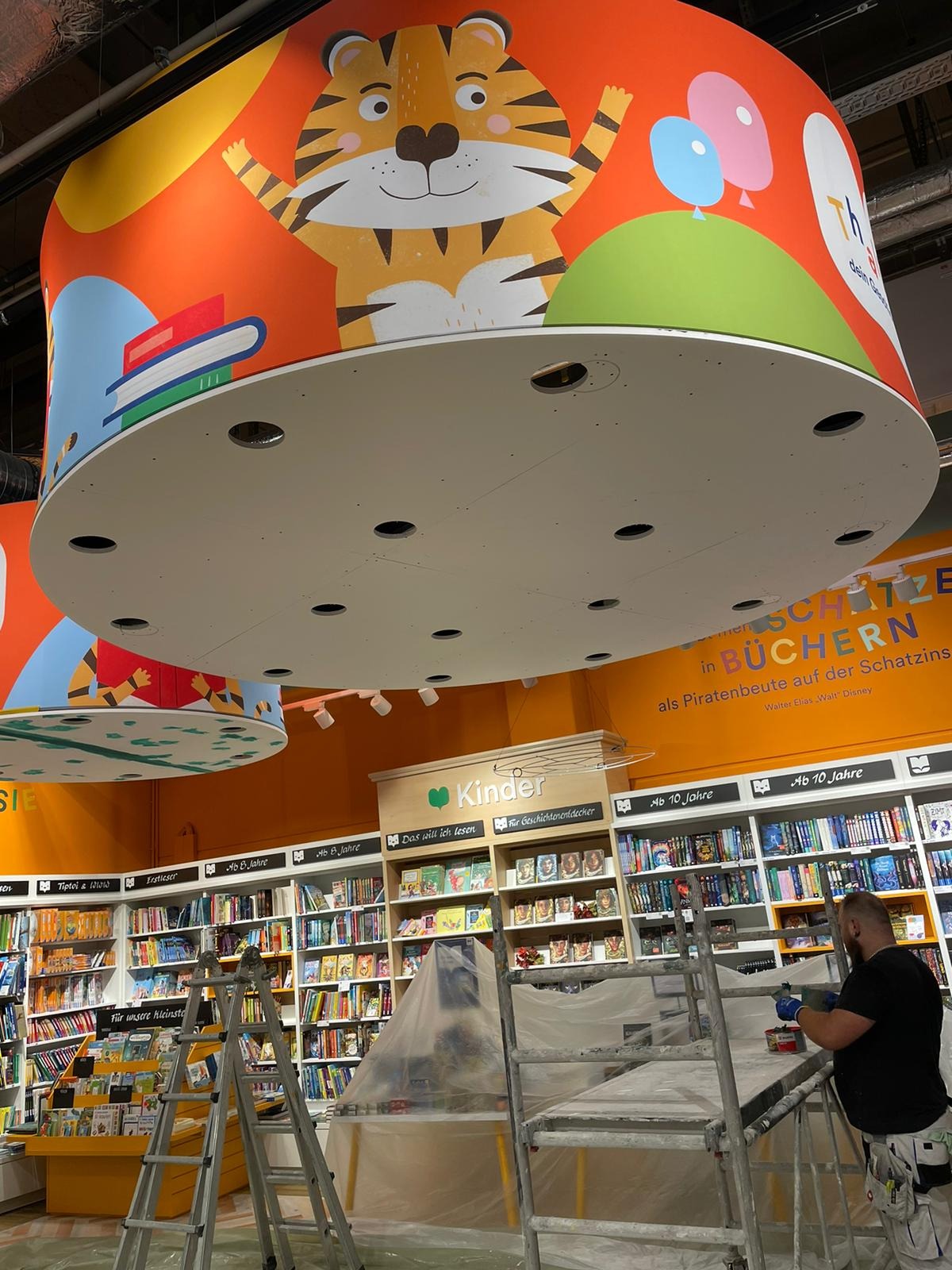 Mężczyzna na rusztowaniu maluje sufit w księgarni dziecięcej, widoczne regały z książkami, kolorowe dekoracje sufitowe z motywami zwierzęcymi i balonami.