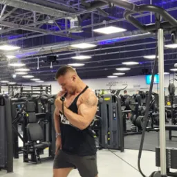 Mężczyzna w czarnej koszulce na ramiączkach ćwiczy biceps na maszynie w siłowni, widoczny tatuaż na ramieniu i zegarek na nadgarstku.