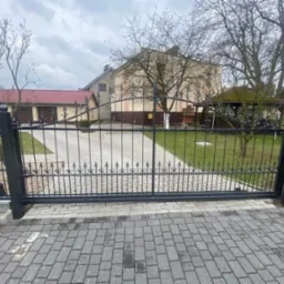 Czarna, metalowa brama wjazdowa z ozdobnymi elementami na posesji z kostki brukowej, widok na budynek gospodarczy i altanę w tle.