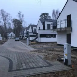 Ulica z kostki brukowej prowadząca do szeregu nowoczesnych domów z balkonami i schodami zewnętrznymi, w tle widoczny samochód i drzewa bez liści, pochmurna pogoda.