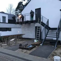 Spawanie metalowej konstrukcji balkonu z schodami zewnętrznymi przy nowoczesnym domu, widoczne iskry, drabina oparta o balustradę, narzędzia na ziemi.
