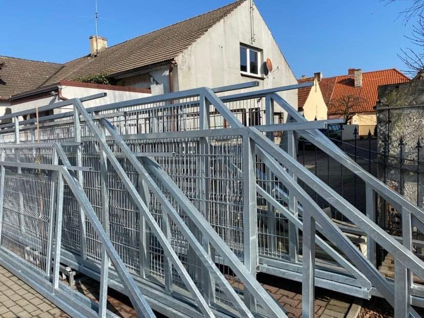 Stos ocynkowanych paneli ogrodzeniowych z siatki z elementami konstrukcyjnymi, przygotowanych do montażu na tle budynków mieszkalnych w słoneczny dzień.