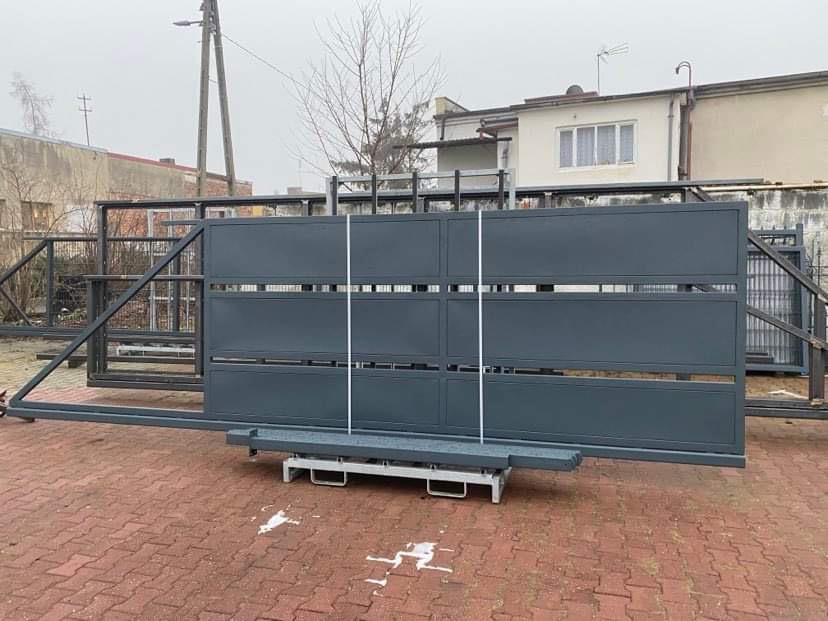 Przygotowana do montażu przesuwna brama wjazdowa w kolorze grafitowym, widoczna na ceglanym bruku przed budynkiem, z elementami konstrukcyjnymi w tle.