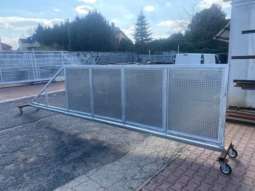 Przesuwna metalowa brama z perforowanymi panelami, zamontowana na wózkach z kółkami, widoczna na zewnątrz, z budynkami i zielenią w tle.