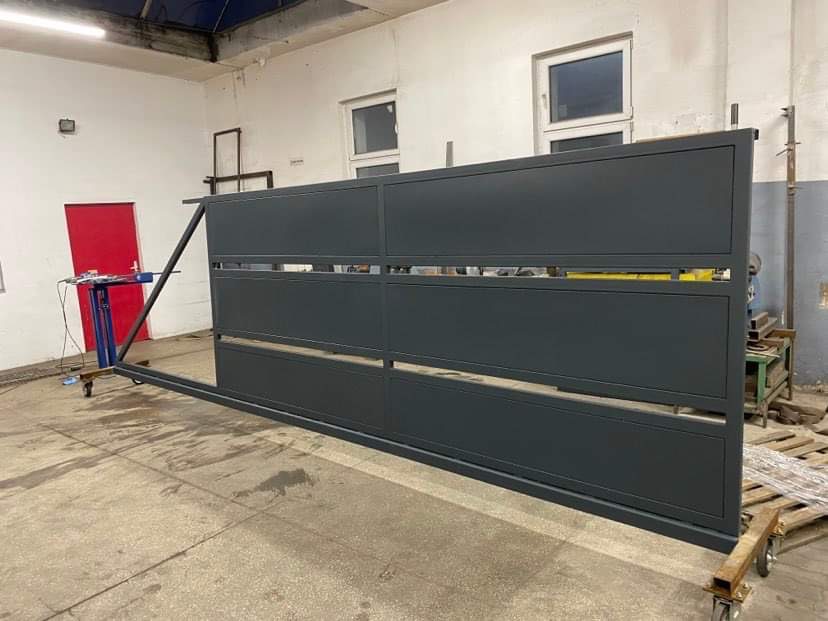 Szara, przesuwna brama wjazdowa w trakcie produkcji, widoczna w warsztacie z narzędziami i paletami, na kółkach transportowych.