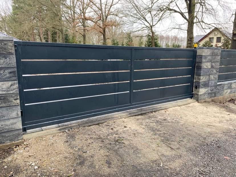 Nowoczesna, przesuwna brama wjazdowa w kolorze antracytowym z poziomymi, srebrnymi akcentami, osadzona pomiędzy słupkami z szarej kostki łupanej, widok z zewnątrz posesji.