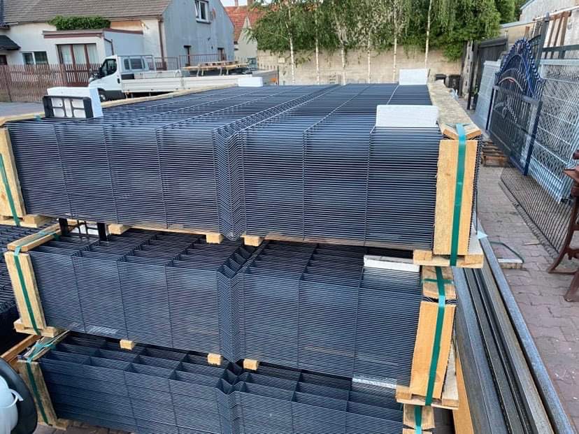 Stos paneli ogrodzeniowych w kolorze grafitowym, spiętych drewnianymi belkami i zieloną taśmą, przygotowanych do transportu na tle zabudowań.