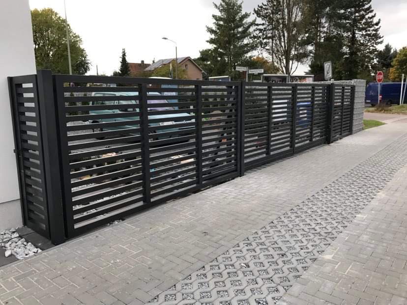 Nowoczesna, pozioma, metalowa brama wjazdowa w kolorze antracytowym, z widocznym podjazdem z kostki brukowej i elementami zieleni w tle.