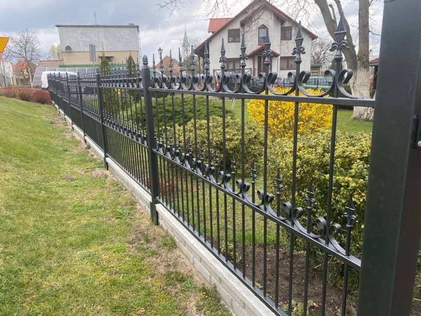 Czarna, metalowa, ozdobna, długa linia ogrodzenia z betonową podmurówką, na tle zielonego trawnika, krzewów i domu.