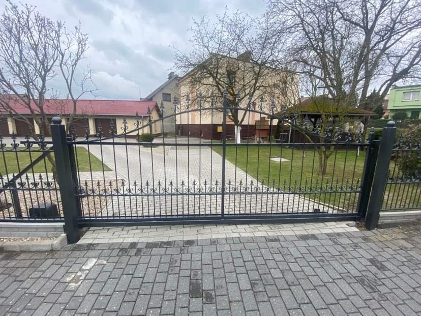 Czarna, metalowa brama wjazdowa z ozdobnymi elementami na posesji z kostki brukowej, widok na budynek gospodarczy i altanę w tle.