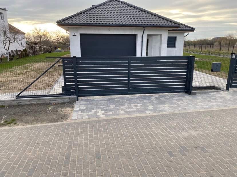 Nowoczesna, przesuwna brama wjazdowa z poziomymi panelami w kolorze antracytowym, zamontowana przed domem z garażem, widoczna siatka ogrodzeniowa w tle.
