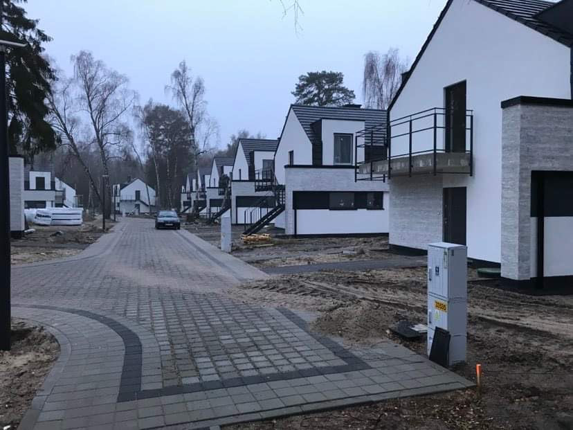 Ulica z kostki brukowej prowadząca do szeregu nowoczesnych domów z balkonami i schodami zewnętrznymi, w tle widoczny samochód i drzewa bez liści, pochmurna pogoda.