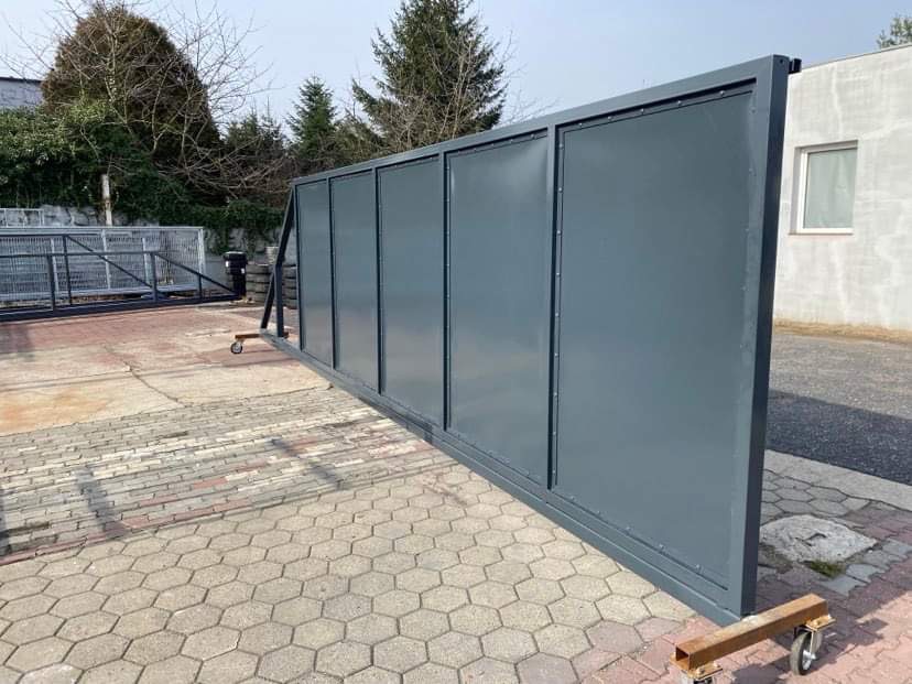 Szara, przesuwna brama wjazdowa z metalowymi panelami, tymczasowo podparta na kółkach, na tle kostki brukowej i budynku.