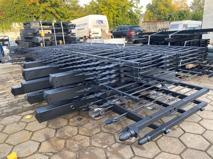 Stos pomalowanych na czarno, metalowych elementów ogrodzenia z dekoracyjnymi grotami, leżących na brukowanej powierzchni, z widocznym w tle samochodem dostawczym firmy.