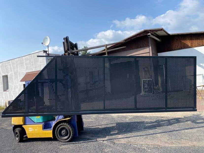 Część ażurowej, metalowej bramy w kolorze grafitowym, transportowana wózkiem widłowym na zewnątrz budynku.