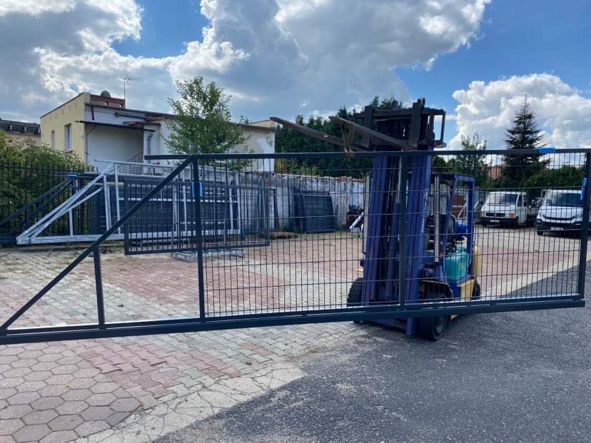 Wózek widłowy transportuje czarną, metalową bramę przesuwną na tle placu z kostki brukowej, częściowo widoczne elementy ogrodzenia i budynek.