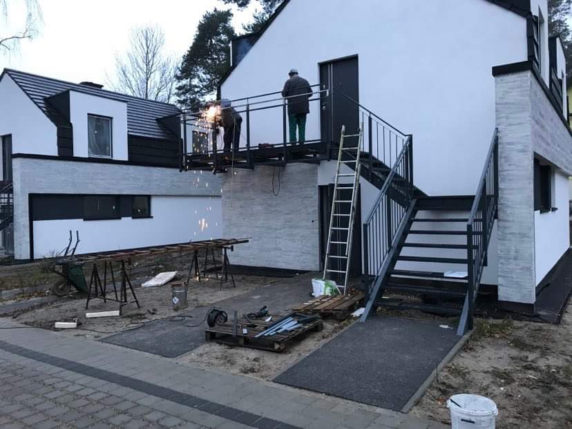 Spawanie metalowej konstrukcji balkonu z schodami zewnętrznymi przy nowoczesnym domu, widoczne iskry, drabina oparta o balustradę, narzędzia na ziemi.