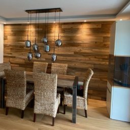 PG- Creative Design - Jadalnia z drewnianą ścianą z desek, stołem i krzesłami z rattanu, oraz lampą wiszącą z szarymi kloszami.