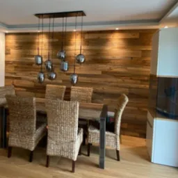 Jadalnia z drewnianą ścianą z desek, stołem i krzesłami z rattanu, oraz lampą wiszącą z szarymi kloszami.