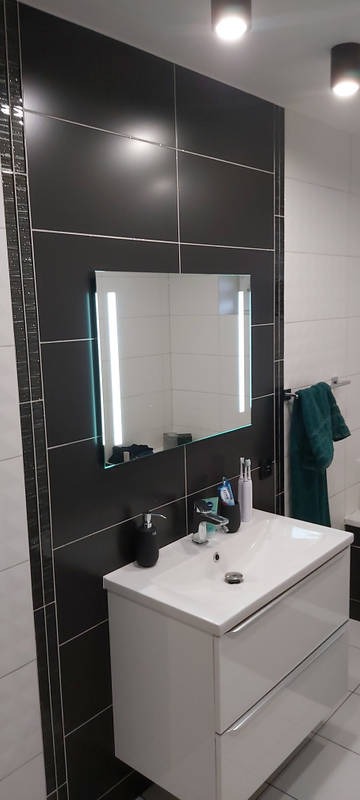 Nowoczesna łazienka z czarno-białymi płytkami, podświetlanym lustrem i białą szafką umywalkową z chromowanymi uchwytami.