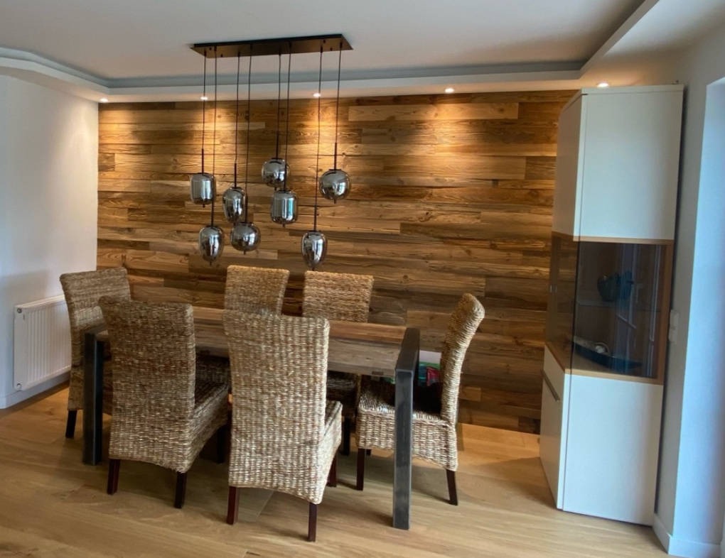 Jadalnia z drewnianą ścianą z desek, stołem i krzesłami z rattanu, oraz lampą wiszącą z szarymi kloszami.