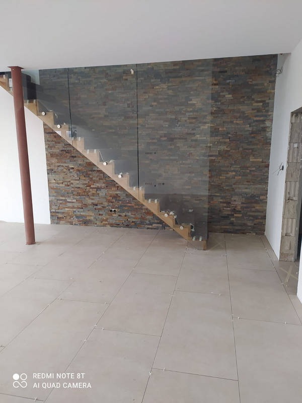 Ściana wykończona kamieniem dekoracyjnym za drewnianymi schodami z szklaną balustradą, widoczne płytki podłogowe przed fugowaniem.