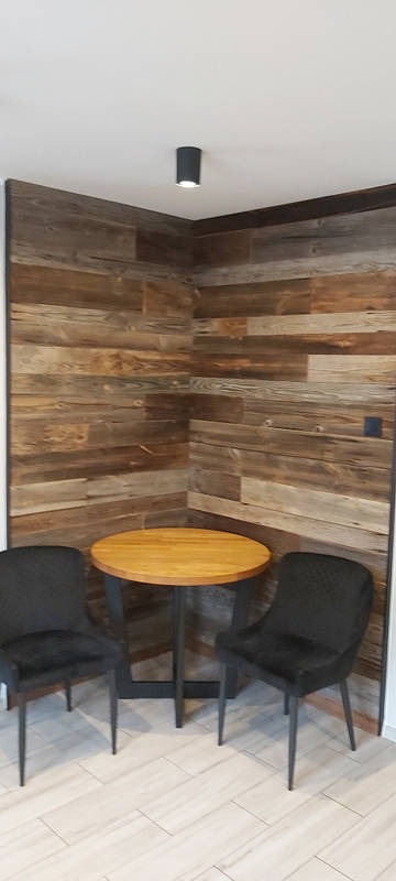 Rustykalna ściana z drewnianych paneli za okrągłym stołem i dwoma czarnymi krzesłami, minimalistyczna lampa sufitowa, jasna podłoga z płytek imitujących drewno.