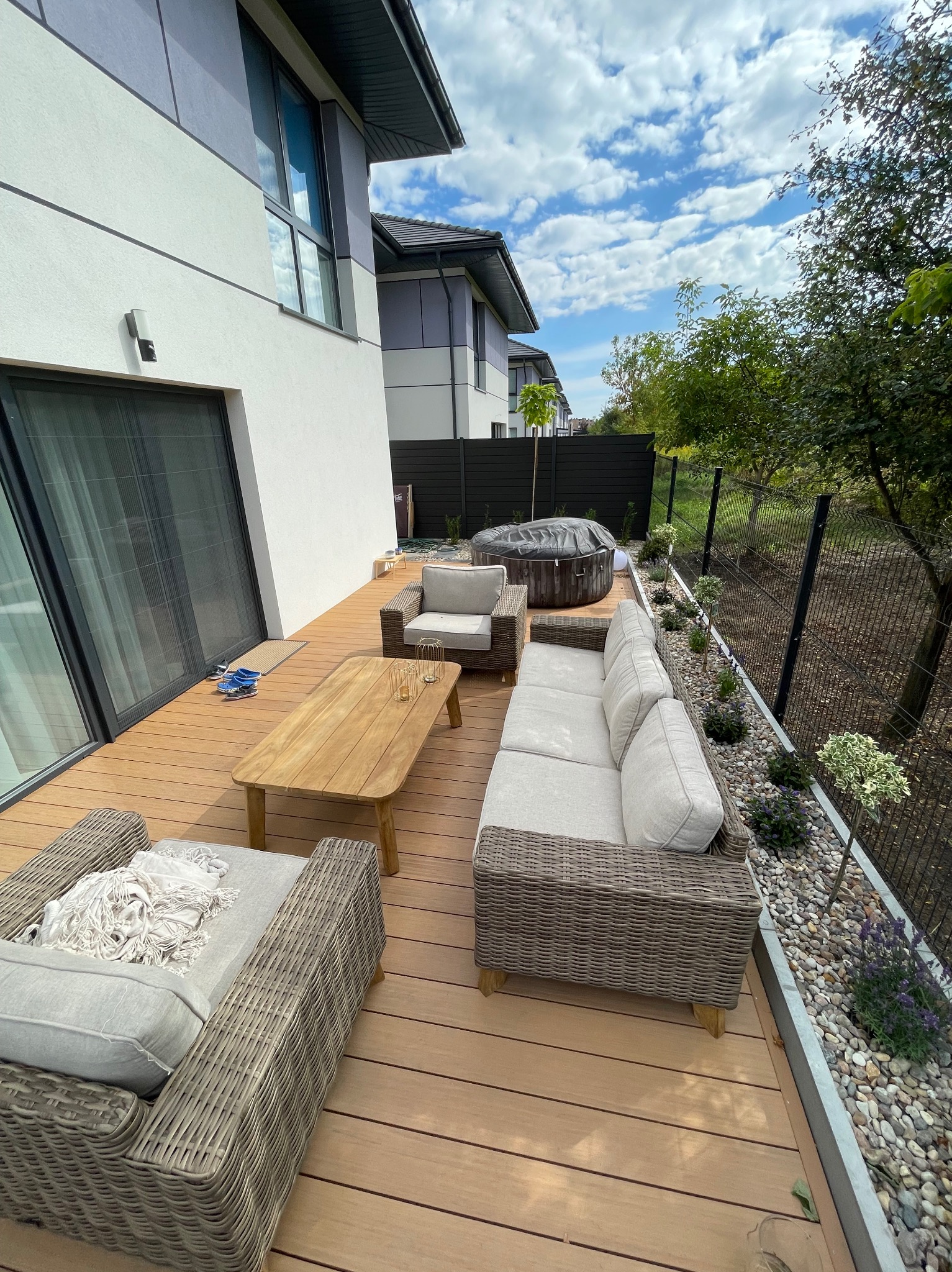 Nowoczesny taras z desek kompozytowych, rattanowe meble wypoczynkowe, jacuzzi i rabata z kamyków oraz roślin, widok z góry, słoneczny dzień.
