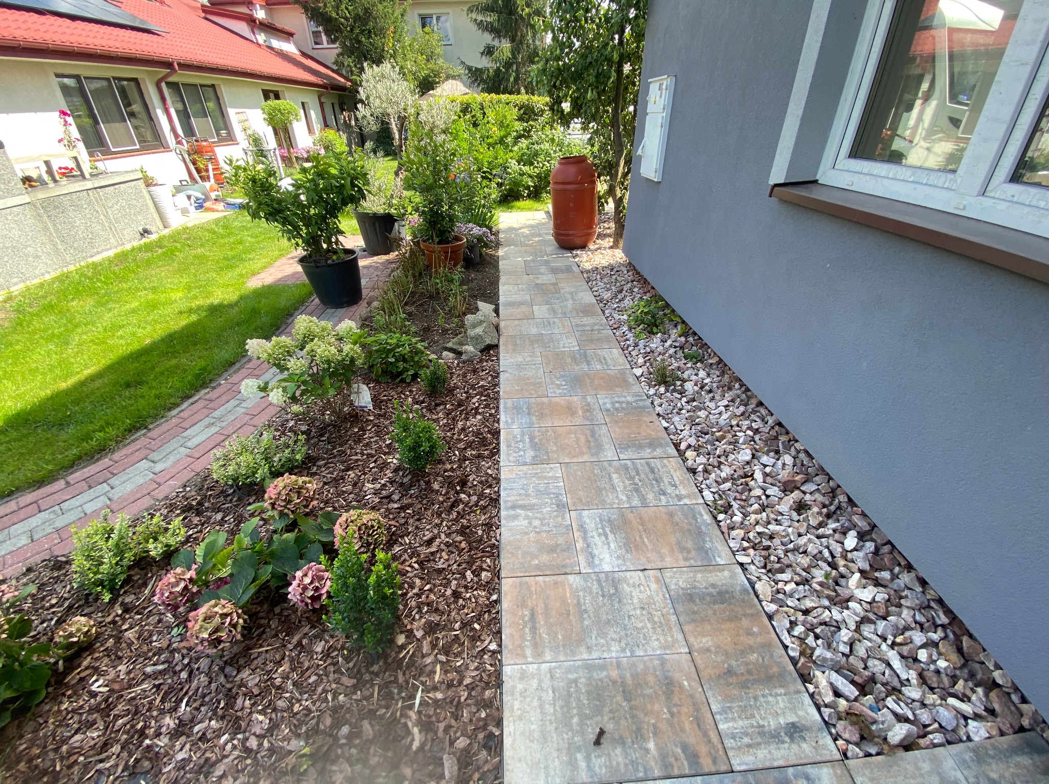 Ułożona ścieżka z kostki brukowej prowadząca wzdłuż domu, obok rabaty z korą, kwiatami i krzewami. Po prawej stronie kamyczki. W tle widoczny budynek i zieleń.