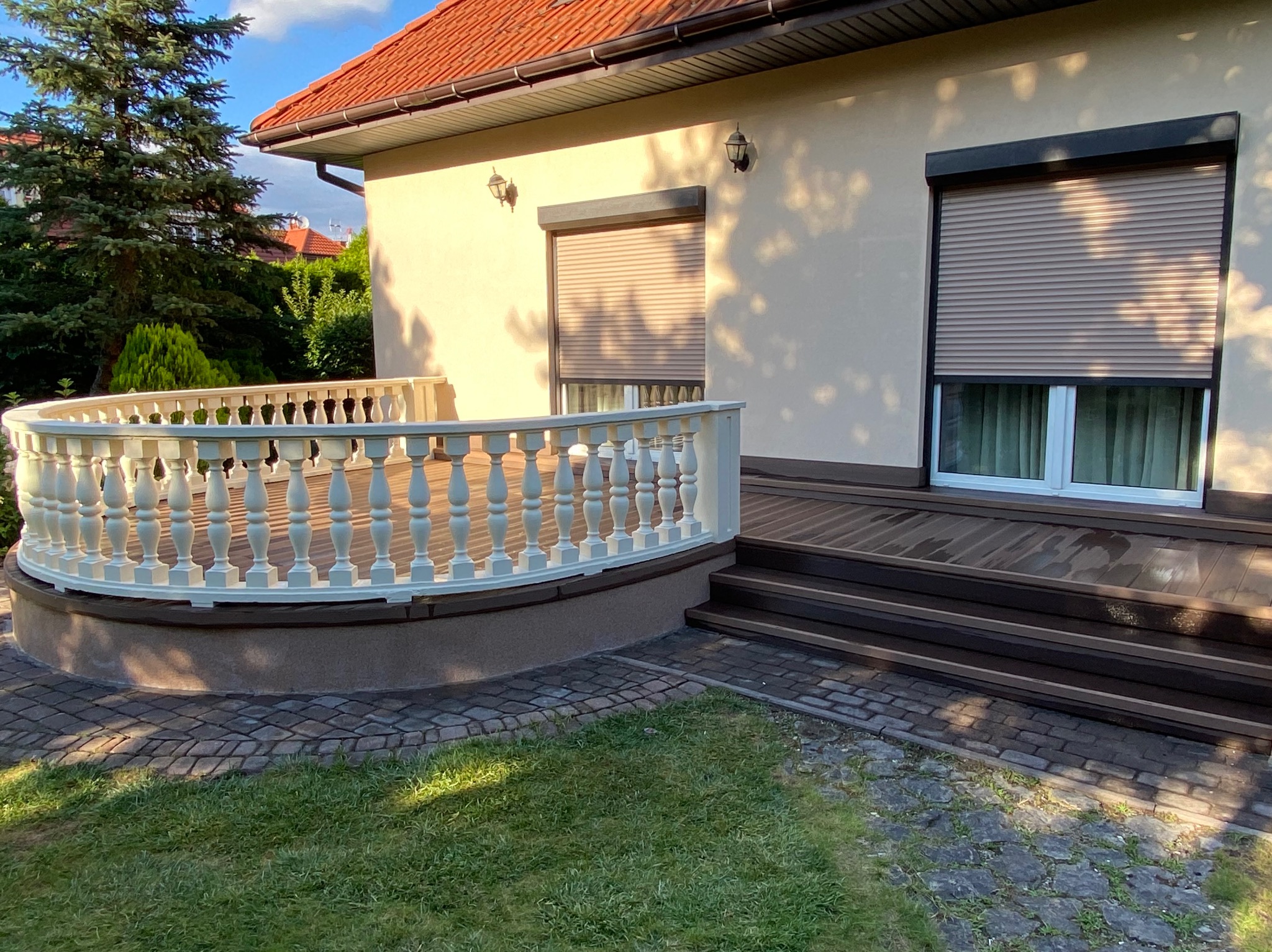 Elegancki taras z drewnianą podłogą i białymi balustradami, przylegający do domu z roletami zewnętrznymi i kostką brukową. Widok na zadbany trawnik i zieleń.