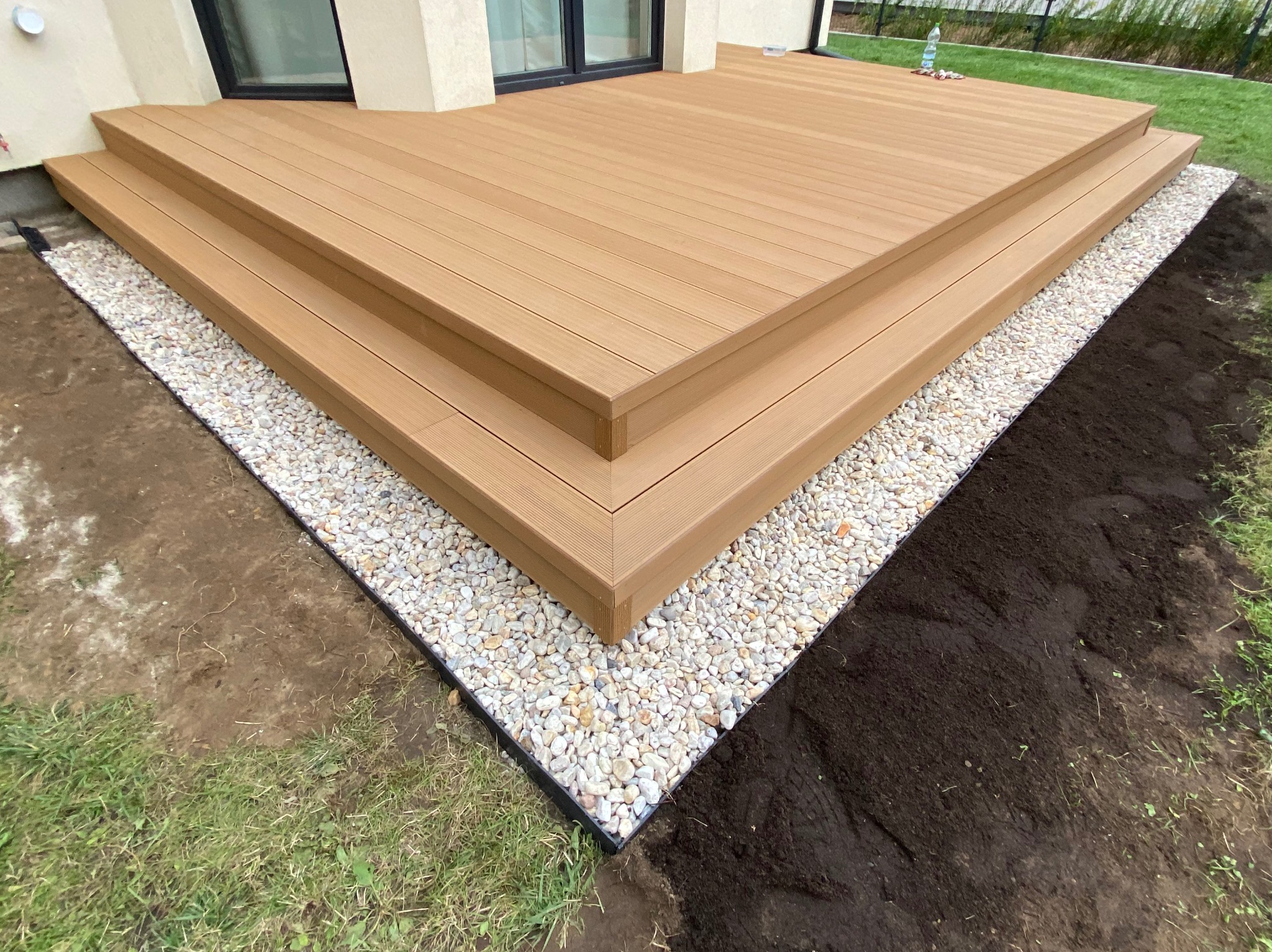 Nowoczesny taras z deski kompozytowej w odcieniach brązu, otoczony białymi kamykami i świeżo przygotowaną ziemią pod nasadzenia, przylegający do jasnej elewacji budynku.