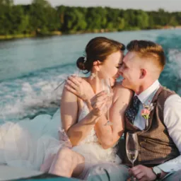 Para młoda świętuje w łodzi motorowej z szampanem, w tle woda i zieleń.