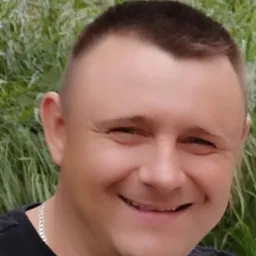 Uśmiechnięty mężczyzna w czarnej koszulce z srebrnym łańcuszkiem na tle zielonych traw.