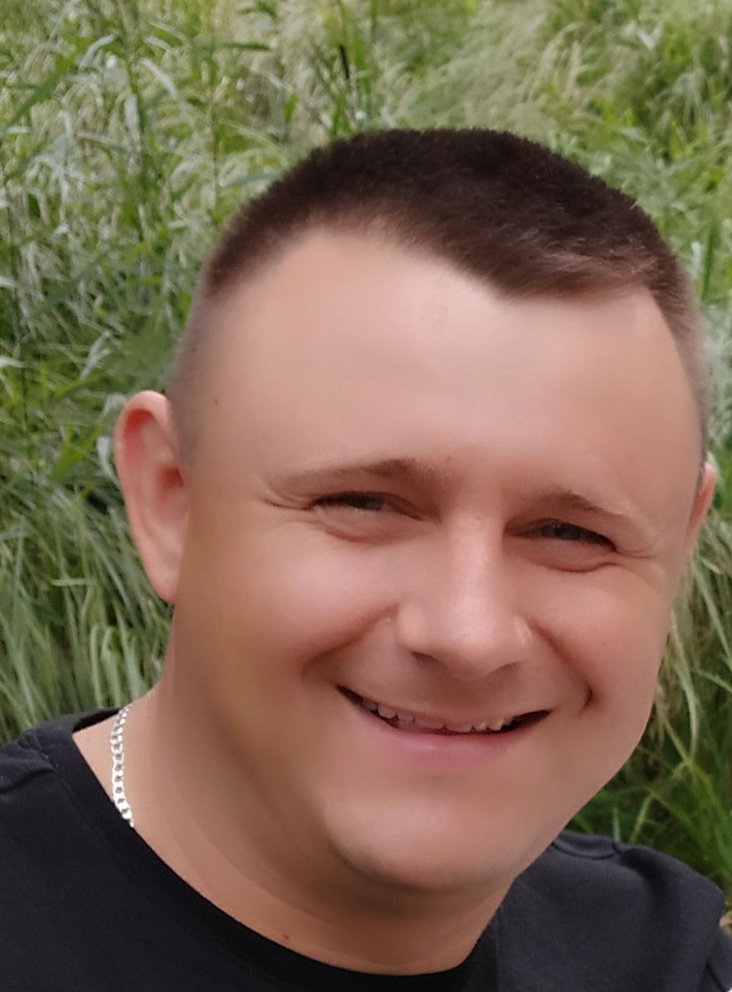 Uśmiechnięty mężczyzna w czarnej koszulce z srebrnym łańcuszkiem na tle zielonych traw.