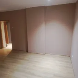 Pomalowane na brązowo ściany w pustym pokoju z panelami podłogowymi, widok na otwarte drzwi do kolejnego pomieszczenia.